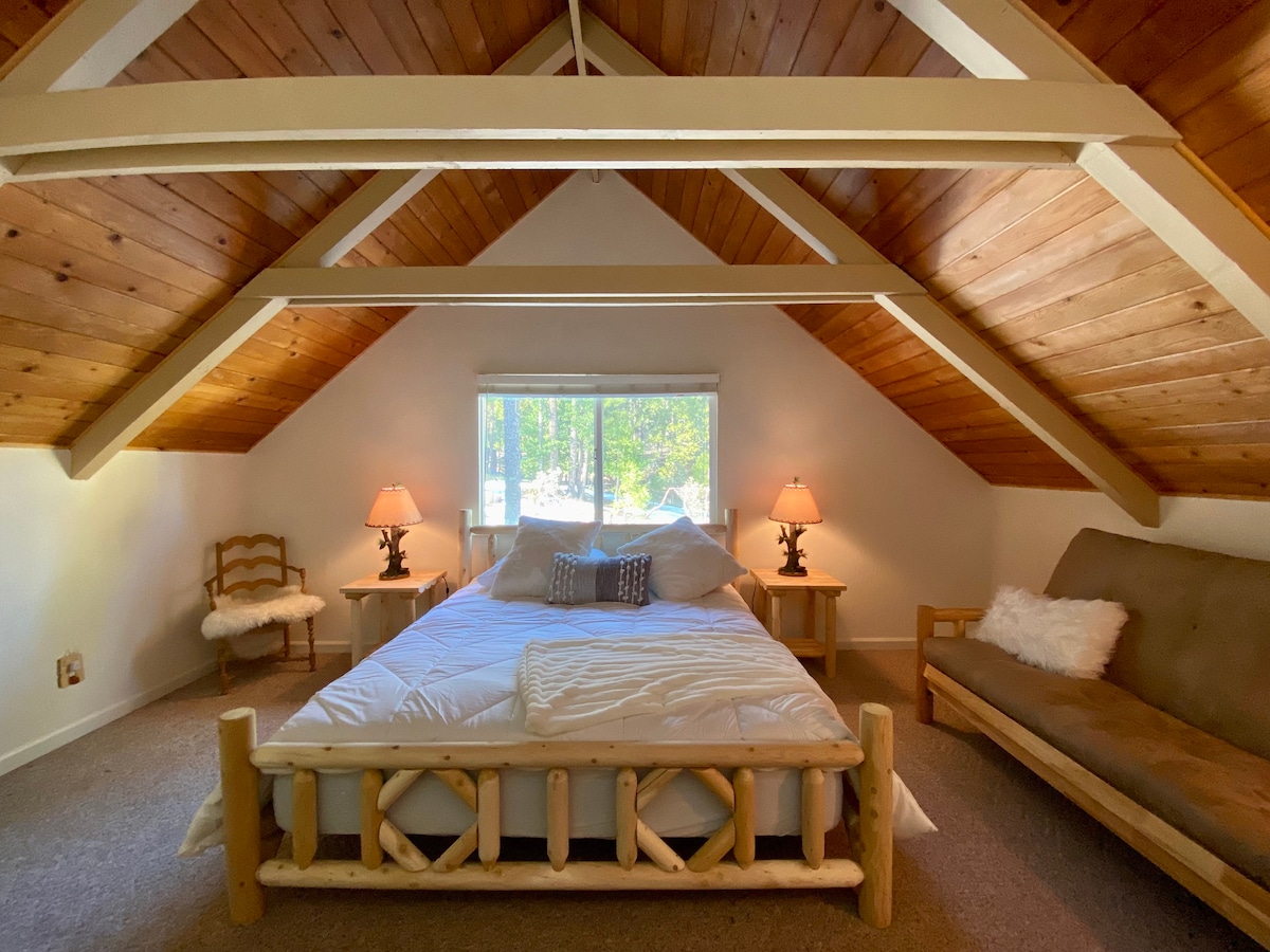 A spacious bedroom features a king bed framed by natural wood accents and a cozy futon. Two lamps provide soft lighting, and a large window allows natural light to illuminate the space, offering views of the surrounding greenery.