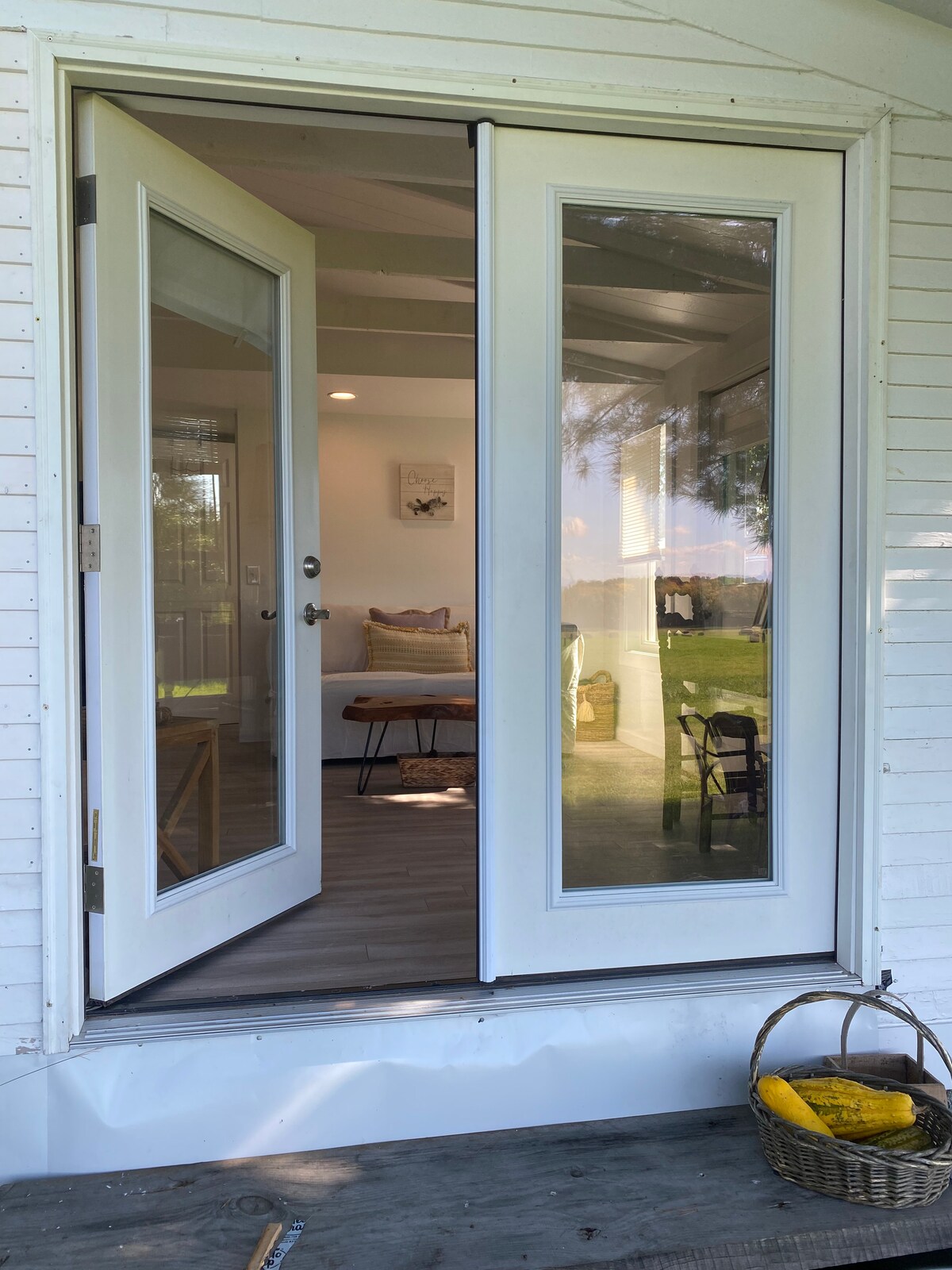 The entrance is framed by double glass doors, revealing glimpses of the interior space. Light filters through the glass, highlighting a welcoming atmosphere. A small wooden table and chairs are visible, along with a basket on the porch floor, suggesting a cozy outdoor area.
