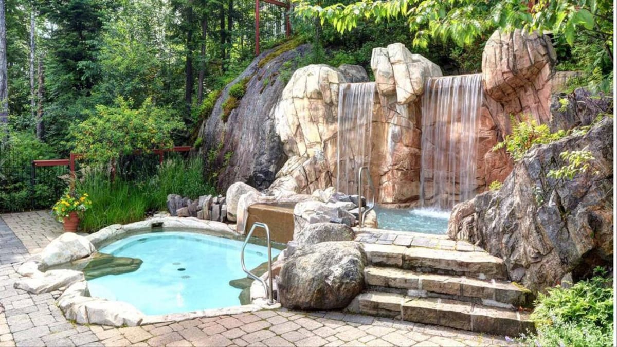 A naturally landscaped outdoor area features a hot tub alongside a tranquil swimming pool. A rock waterfall flows gently over stones, surrounded by dense greenery. Paved stone steps lead down from the surrounding patio, which is adorned with vibrant plants.