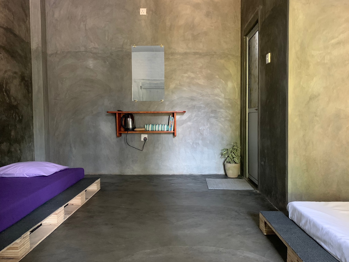A minimalist room is featured with a polished concrete floor and textured gray walls. Two low-profile beds are positioned on wooden pallets, with a shelf displaying a kettle and cups. A small plant adds a touch of greenery in the corner.