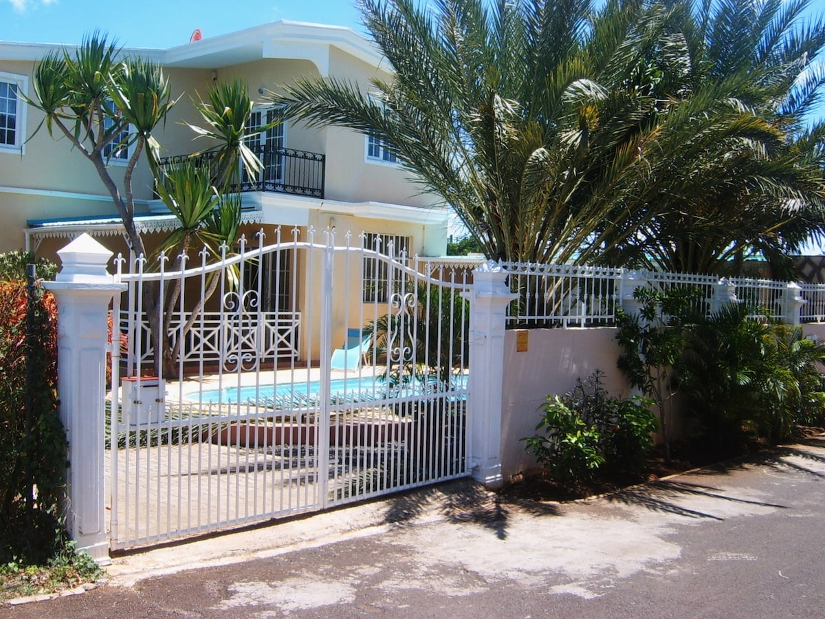 A gated entrance leads to the villa, framed by palm trees and vibrant greenery. The white iron gate stands open, revealing a glimpse of the pool and the inviting facade of the house, surrounded by lush landscaping.