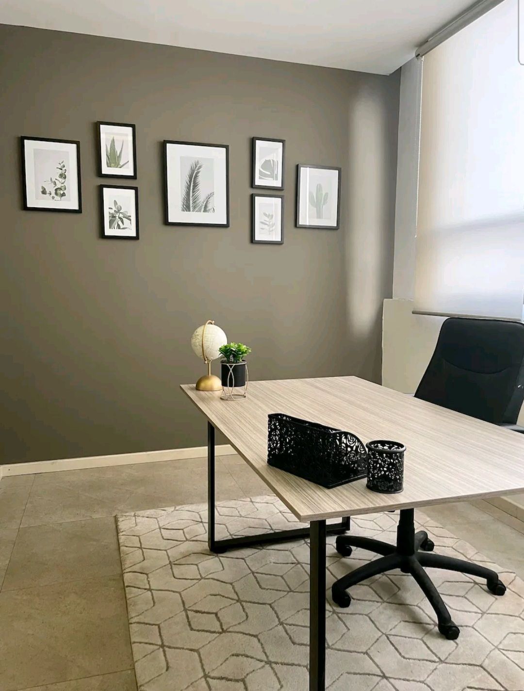 A dedicated workspace features a desk and a black office chair. The desk is adorned with small decorative items and framed art showcasing botanical designs on the walls. Natural light is provided through a window, enhancing the workspace's ambiance.