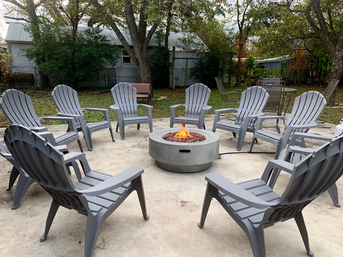 A spacious outdoor area features a circular arrangement of eight gray adirondack chairs surrounding a gas fire pit, with a flame flickering in the center. The setting is shaded by mature oak trees, enhancing the serene atmosphere.
