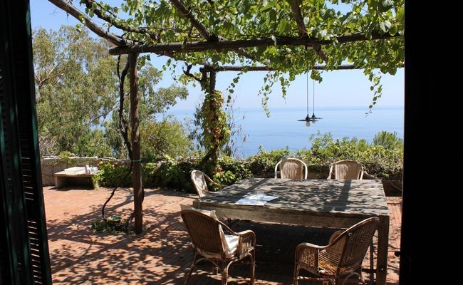 A private terrace is presented with a wooden dining table surrounded by woven chairs, creating an inviting outdoor dining space. Lush greenery and views of the Mediterranean Sea are visible in the background, enhancing the serene environment.