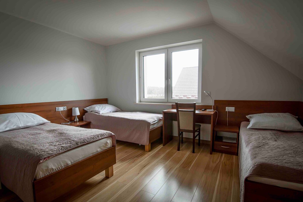A simple room features three beds with neutral bedding, arranged to provide ample space. A desk with a chair is positioned near a bright window, allowing natural light to illuminate the area. Wooden flooring adds warmth to the overall appearance.
