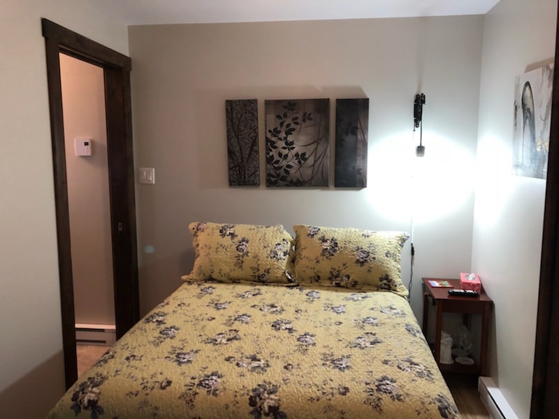 A cozy sleeping area is highlighted by a queen size bed dressed in a floral yellow comforter. Three framed artwork pieces adorn the wall above the bed. A bedside table with a light and decorative items complements the warm color palette of the room.