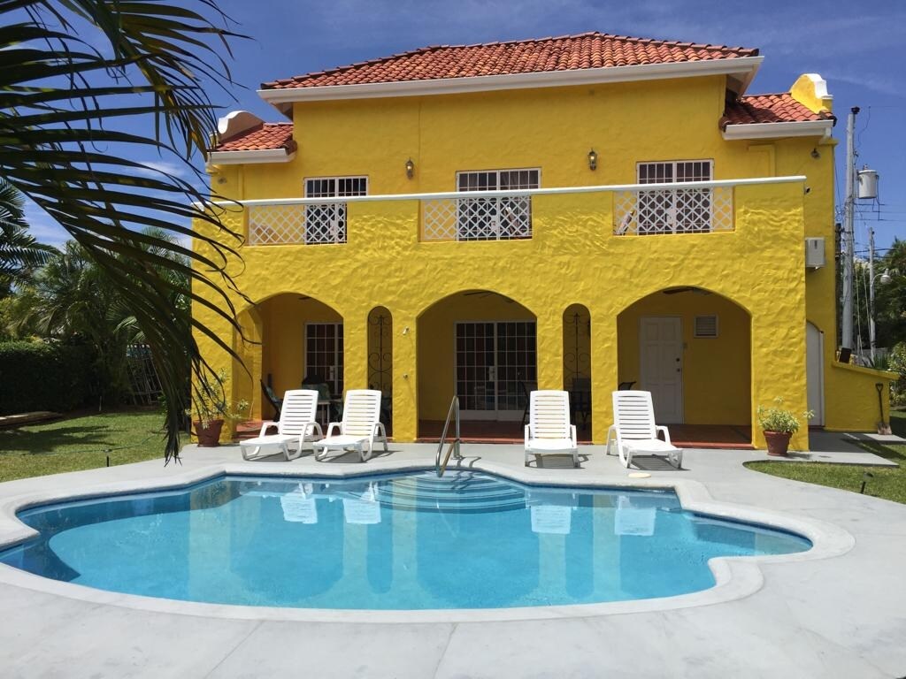 A vibrant yellow villa features a swimming pool surrounded by four lounge chairs. The structure has arched entrances on the ground floor and several windows on the upper level, providing natural light. Lush greenery is visible in the background, contributing to the tropical setting.