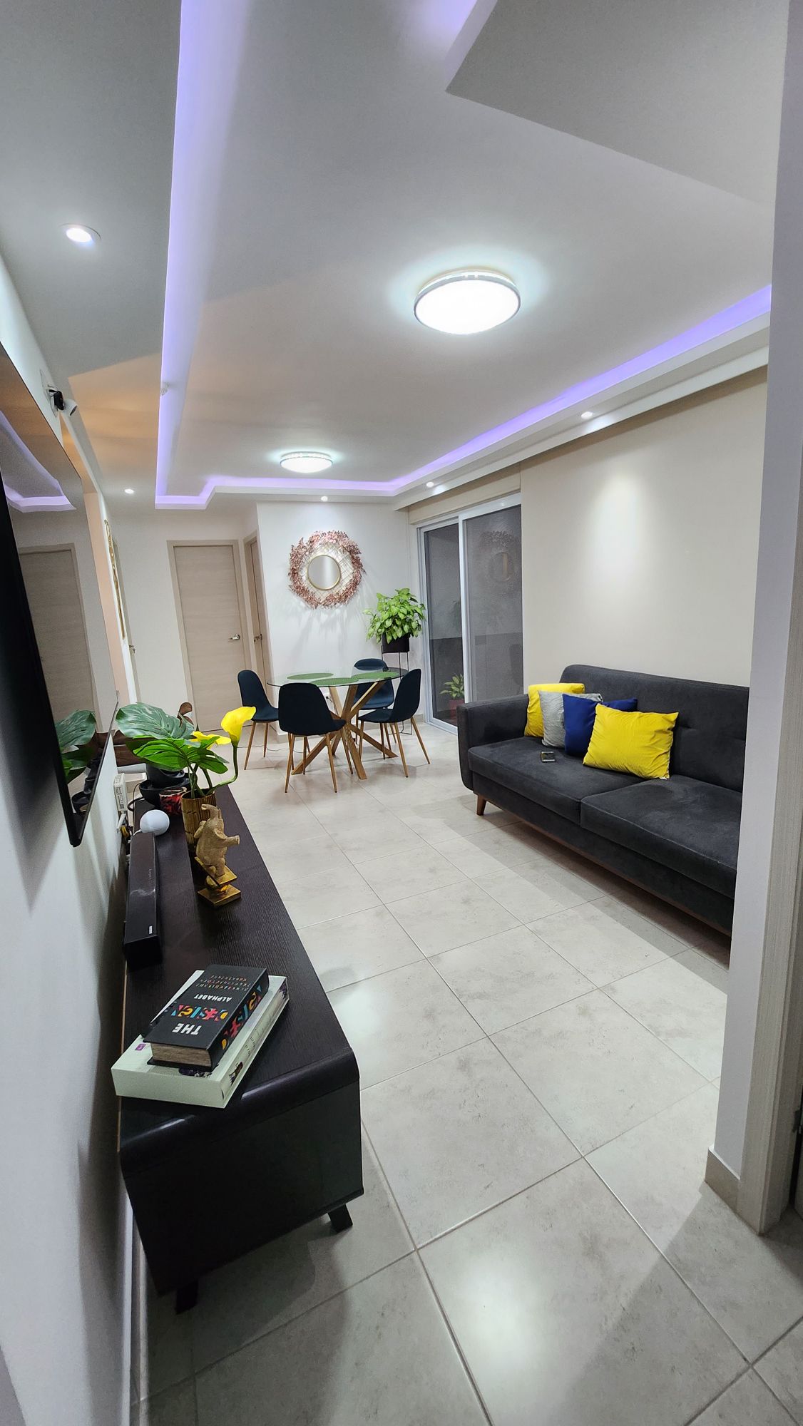 A well-lit living area showcases a dark sofa adorned with yellow accent pillows. A sleek coffee table holds a book, while a dining set for four is positioned nearby. A mirror and decorative plant add depth to the space, complemented by soft ceiling lighting.