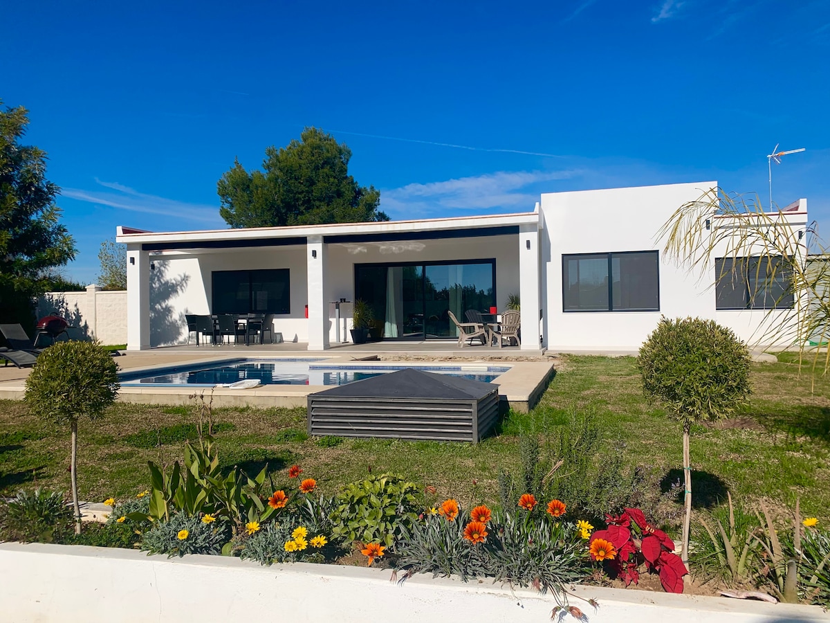 The villa features a modern design complemented by large windows, allowing natural light to fill the interior. A private saltwater pool is centered within the garden, surrounded by green grass and colorful flowers, enhancing the outdoor space. Lounge chairs and a dining area can be seen by the pool.