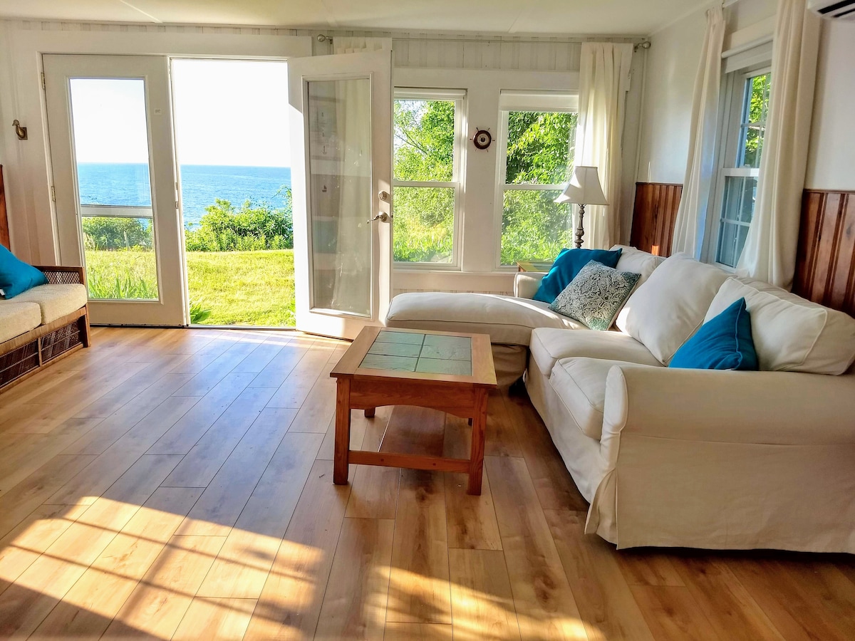 A bright living area features large glass doors opening to an expansive view of Cape Cod Bay. Light wood flooring enhances the warmth of the space, furnished with a comfortable white sofa adorned with blue accent pillows and a wooden coffee table.