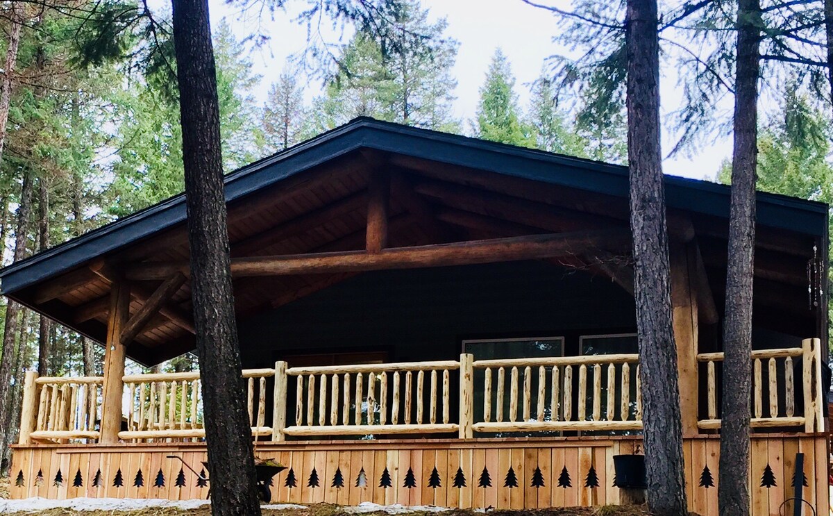 A cabin exterior is highlighted by a large, covered porch with a rustic wooden railing. Tall trees surround the property, providing a natural backdrop and shade. The sloped roof of the cabin is crafted with logs, showcasing a cozy, inviting design.