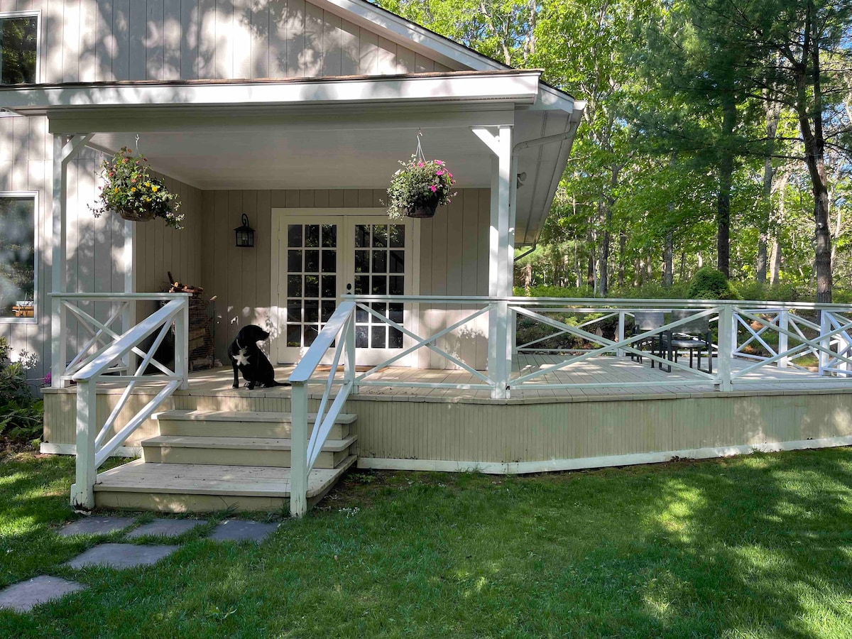 A spacious wooden deck is presented, surrounded by a grassy area. Flower baskets hang attractively from the railing, while a black dog sits on the steps leading down from the deck. French doors add a welcoming touch, connecting the outdoor space to the interior.