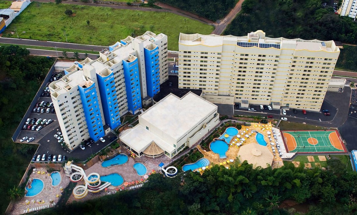 An aerial view showcases the Golden Dolphin Grand Hotel complex, featuring multiple buildings with blue accents. Various natural pools are visible, surrounded by sun loungers and landscaped areas. A nearby recreational court can be seen, adding to the vibrant atmosphere of relaxation and leisure.
