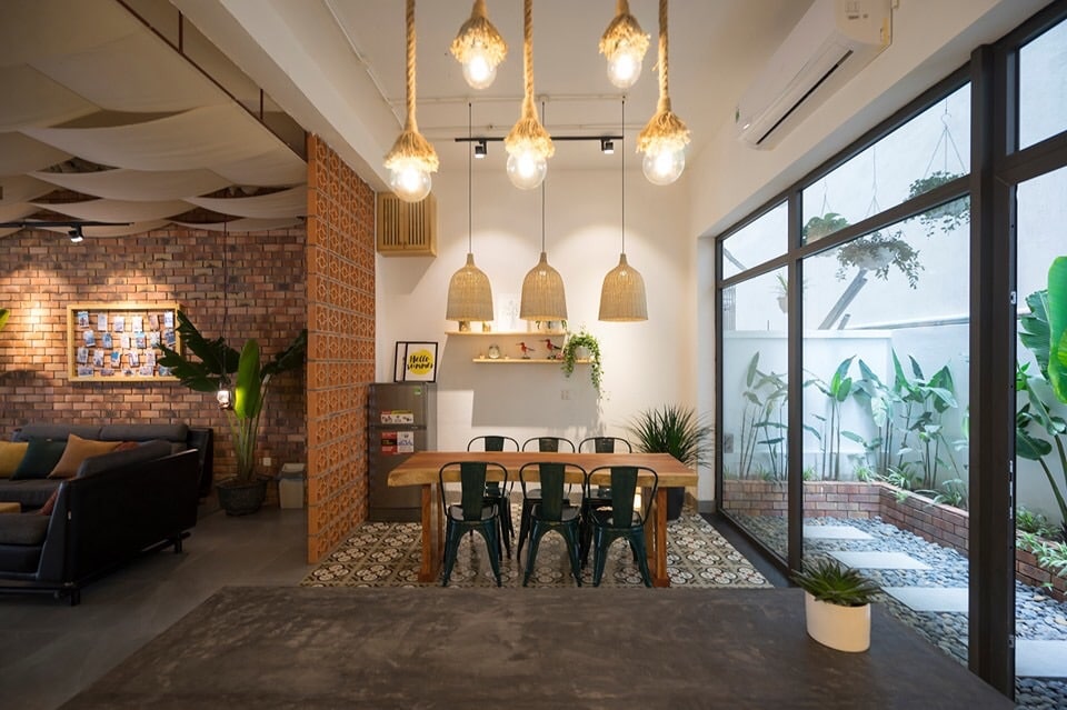 An inviting dining area is highlighted by a wooden table surrounded by green chairs. Three pendant lights made of natural materials hang overhead. Large glass doors offer views of an outdoor space filled with greenery, while patterned tiles add texture to the floor.