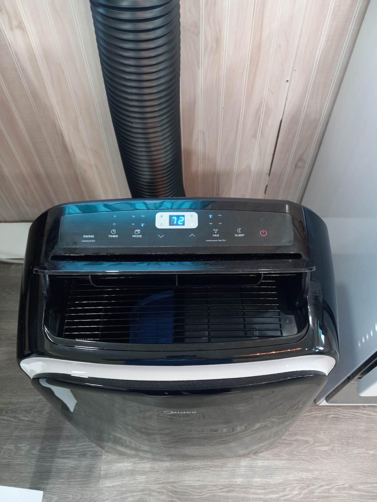 A portable air conditioning unit is shown with a digital control panel featuring various buttons and a temperature display indicating 72 degrees. The unit has a sleek black exterior, and a ventilation hose extends upward, connecting to the wall, while a nearby white refrigerator is partially visible.