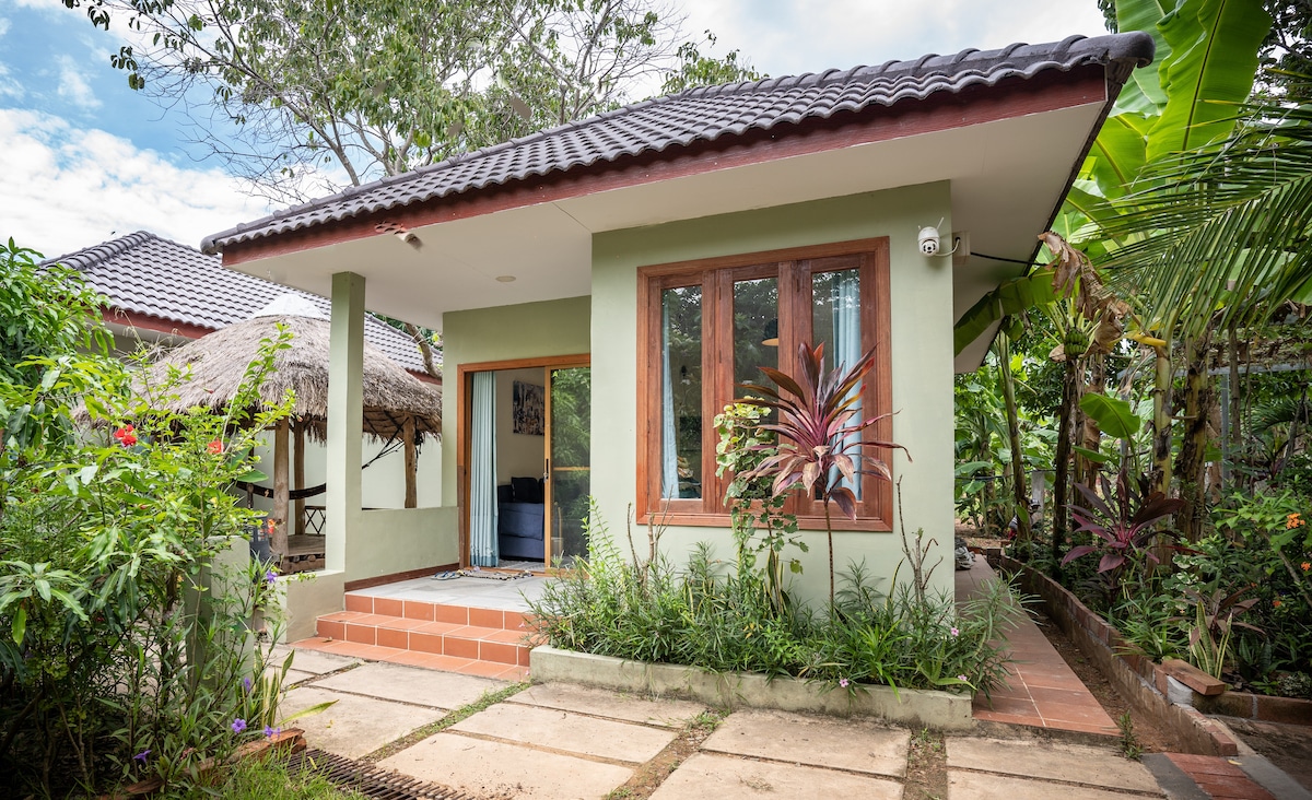 The exterior of a charming bungalow is set amidst lush greenery. A welcoming porch area features a small set of stairs leading to a large front window, which allows natural light to fill the space. Surrounding tropical plants create a serene environment.