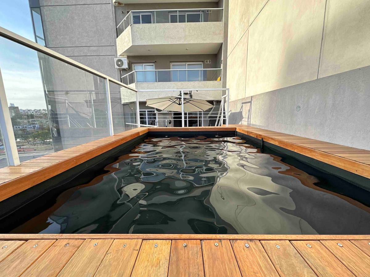 A private heated pool is surrounded by wooden decking, offering a space for relaxation. The pool's sleek design reflects the clear sky, while a view of the modern building's exterior and balcony can be seen in the background.