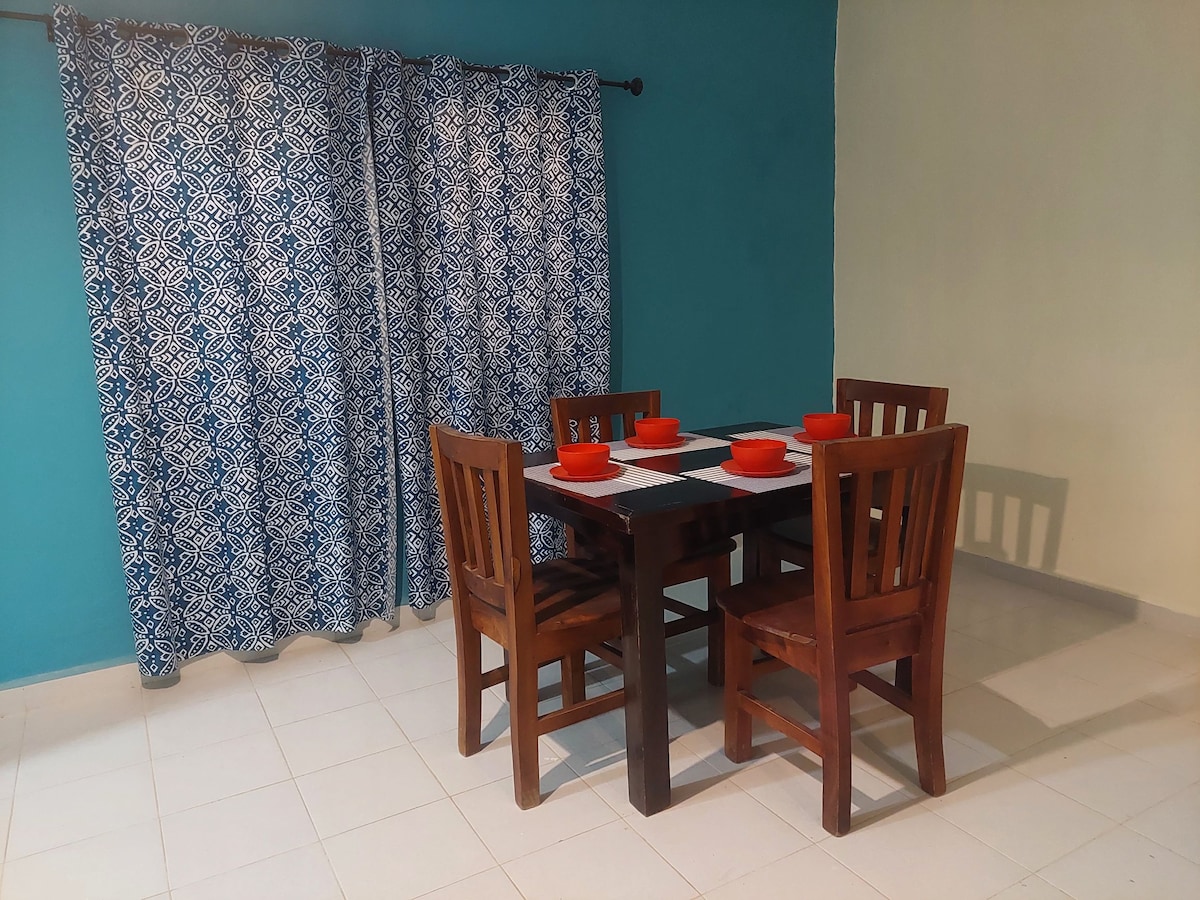 A dining area features a dark wooden table set for four, complemented by matching chairs. Bright red plates are arranged on a black and white table runner. A blue patterned curtain hangs in front of a window, creating a colorful contrast in the spacious room.