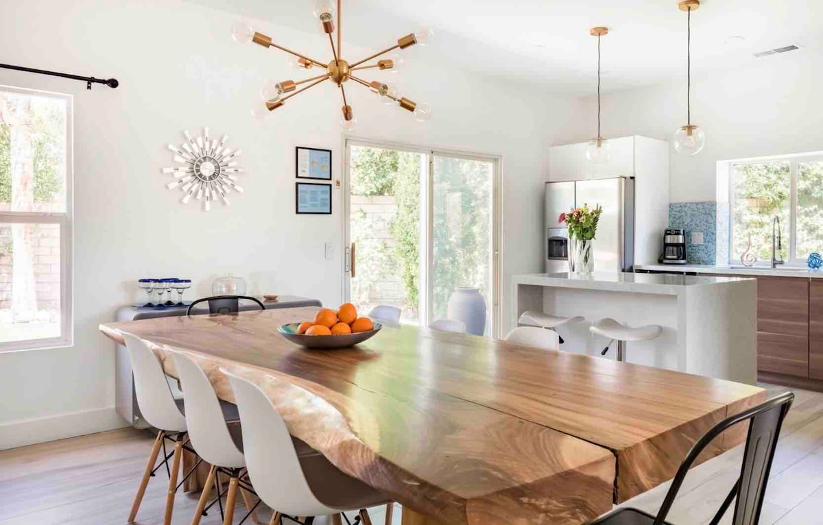 A spacious dining area features a large wooden table with a natural edge, surrounded by a mix of white and black chairs. Bright light enters through large windows, illuminating the space. A bowl of oranges is centrally placed on the table, enhancing the fresh atmosphere.