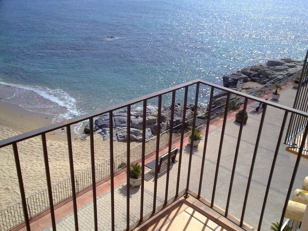 A balcony railing frames a view overlooking the tranquil sea. Gentle waves lap against the sandy beach, while sunlight sparkles on the water surface. Nearby benches and coastal rocks are visible, creating a serene outdoor setting.