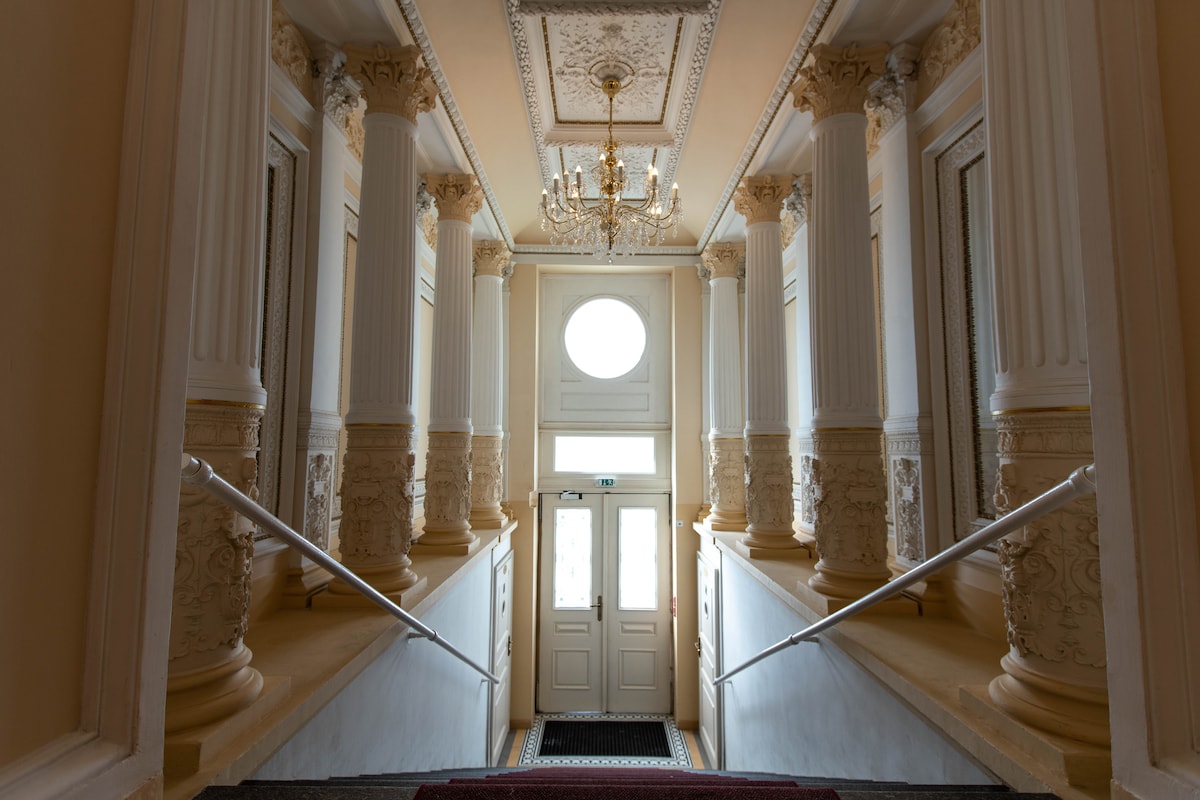 An elegant entryway is framed by tall, decorative columns and features a grand chandelier hanging from the ornate ceiling. Natural light enters through a circular window above the double doors, illuminating the staircase leading down toward the main entrance.