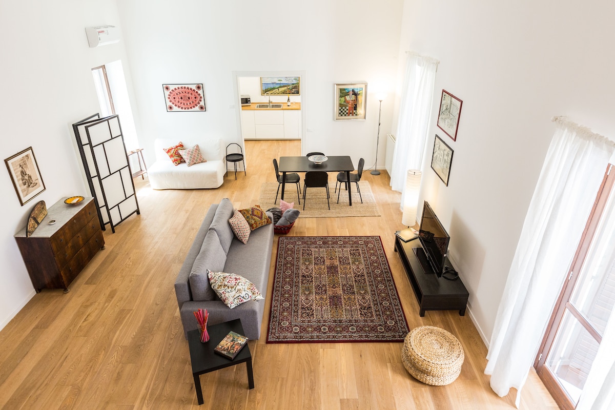 A spacious living area features a comfortable gray sofa adorned with decorative cushions, a wooden sideboard, and a low table. An area rug adds warmth to the wood floor. Light streams through large windows, while artwork decorates the walls, enhancing the inviting ambiance.