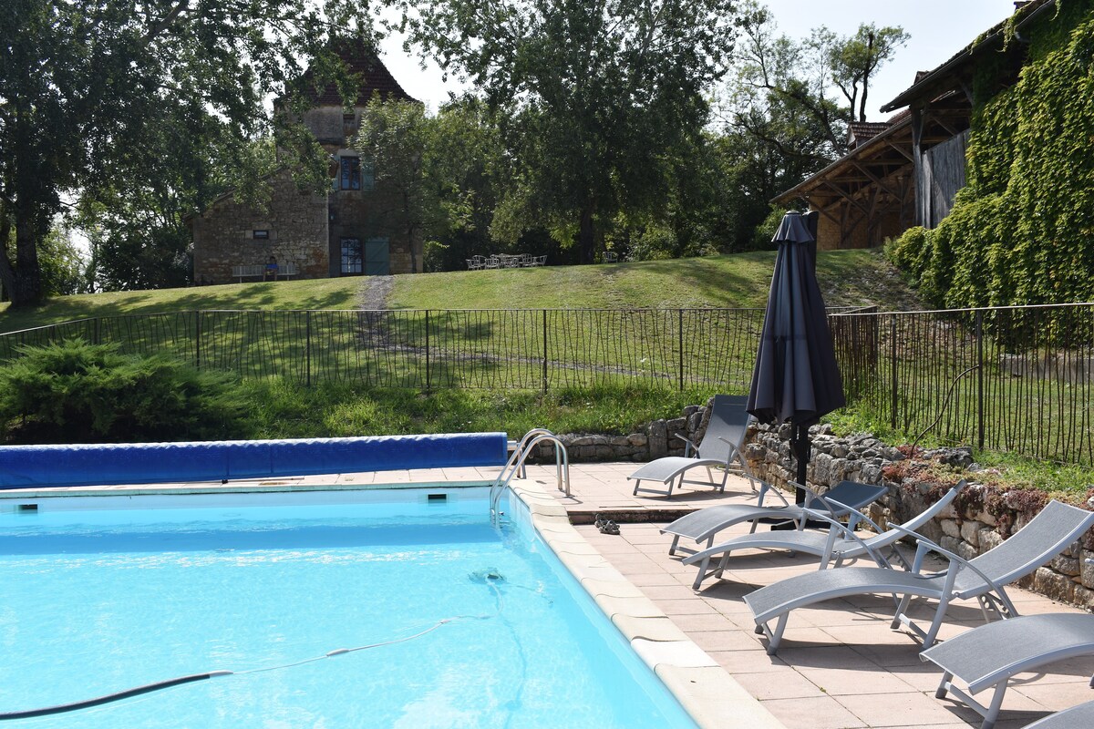 A private swimming pool is surrounded by a paved area featuring sun loungers and an umbrella. The verdant landscape rises gently behind the pool, showcasing a historic building and trees that provide shade and tranquility.