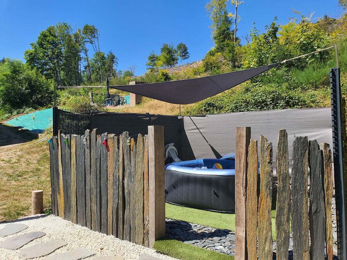A shaded outdoor area features a portable hot tub surrounded by natural greenery. A wooden fence adds privacy, while a pathway of small stones leads into the space. Sunlight filters through the trees, contributing to a serene atmosphere.