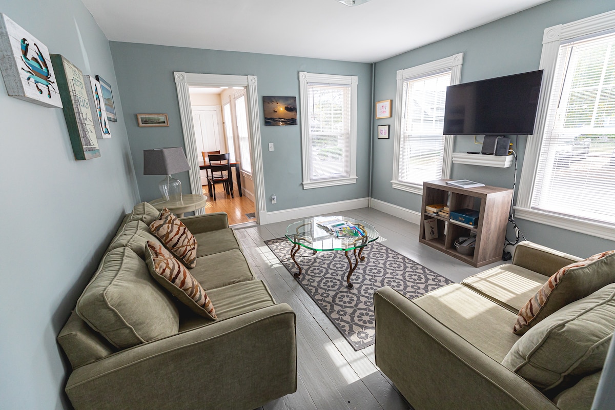 A bright living area features two comfortable sofas, each adorned with patterned cushions. A glass coffee table is centered on a decorative rug. Sunlight streams through multiple windows, illuminating the space, which also includes a television mounted on the wall.