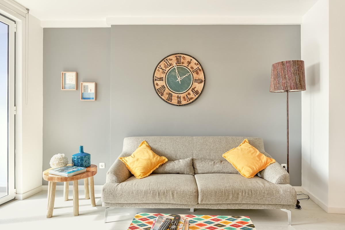 A light-filled living area features a comfortable gray sofa adorned with bright yellow cushions. A round wooden side table displays decorative items, and a colorful coffee table adds a lively touch. A large wooden clock and a standing lamp enhance the stylish decor.