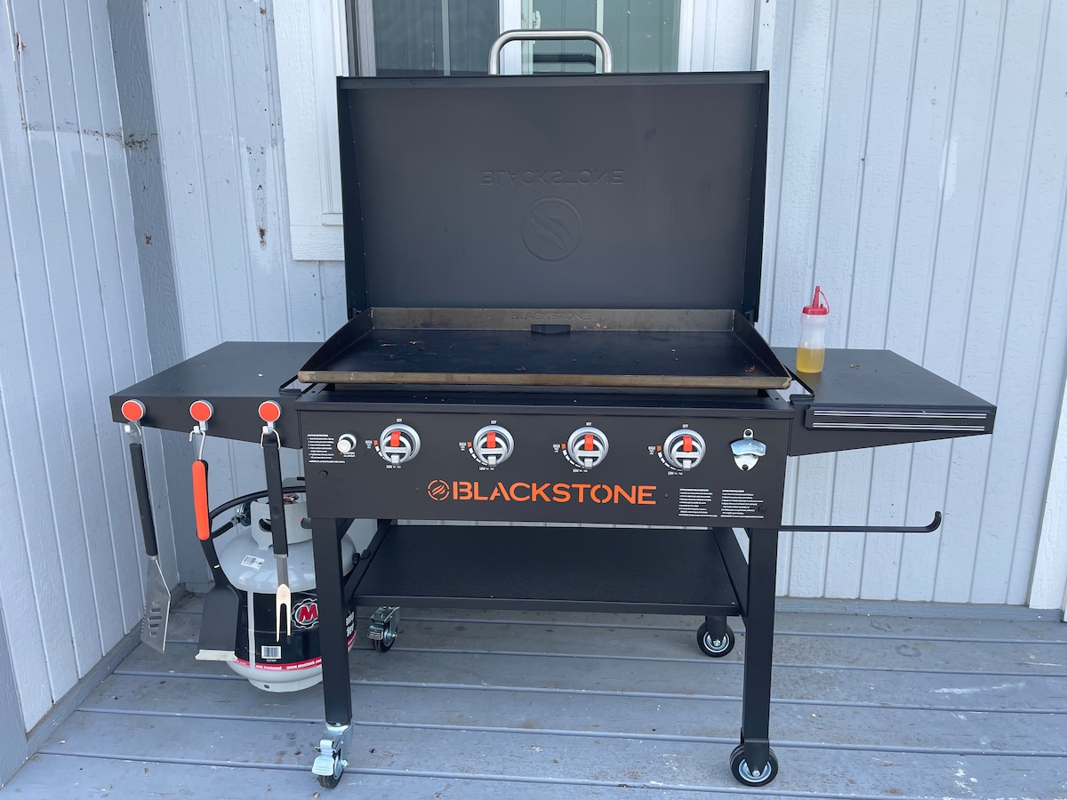 A 36" Blackstone Griddle is displayed on a wooden deck. The griddle features multiple burners with knobs and a bottle of oil placed nearby. The griddle is positioned against a light-colored wooden wall, providing a functional outdoor cooking space.