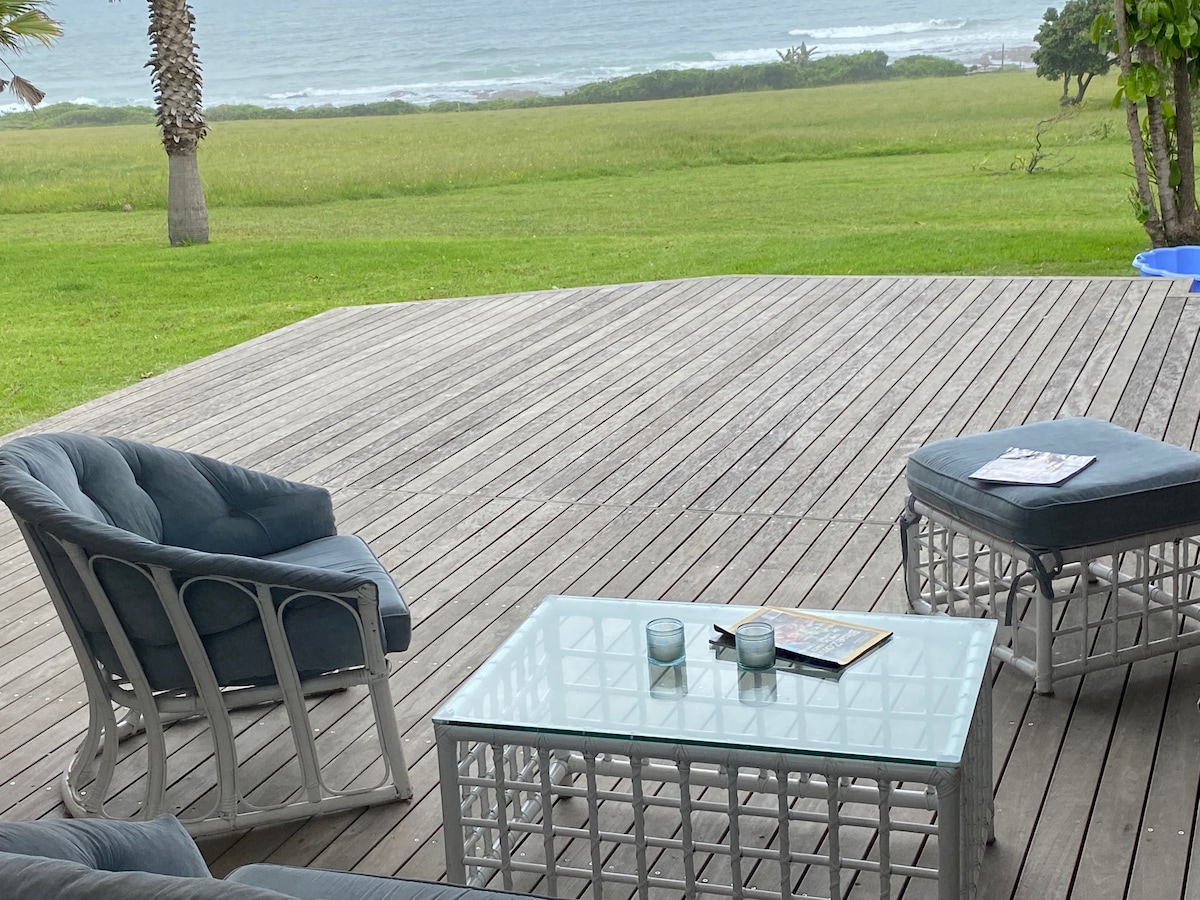 A patio area is depicted, featuring a glass table and two cushioned chairs. A small ottoman is positioned beside the table. Lush green grass extends in the foreground, while the ocean is visible in the background, enhancing the open and relaxing setting.