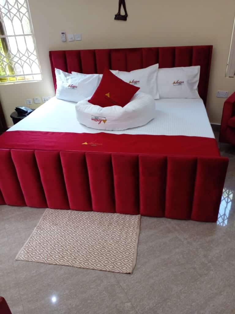 A spacious bed is framed by a deep red upholstered headboard. The bed is adorned with white and red accent pillows, including a round pillow at the center. A light-colored area rug is placed in front of the bed, complementing the polished floor.