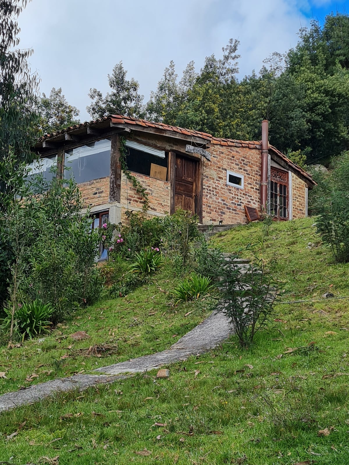 A charming cabin is nestled on a grassy hill, surrounded by lush greenery and trees. The exterior features a mix of stone and brick with large windows that allow natural light inside. A stone pathway leads to the entrance, enhancing the inviting rural setting.