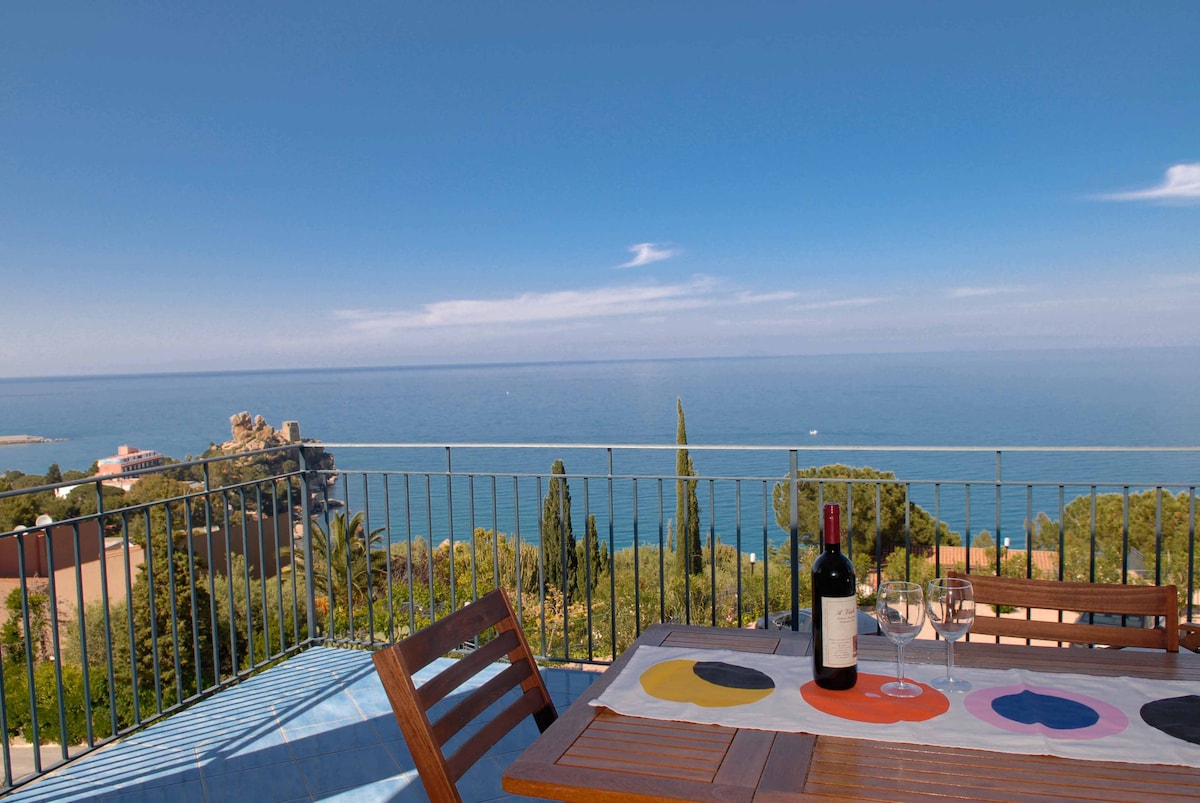 A dining table set for two is positioned on a terrace, overlooking a serene blue sea. A bottle of wine and glasses are placed on the table, with vibrant placemats adding a splash of color. The surrounding landscape features lush greenery and distant rock formations.