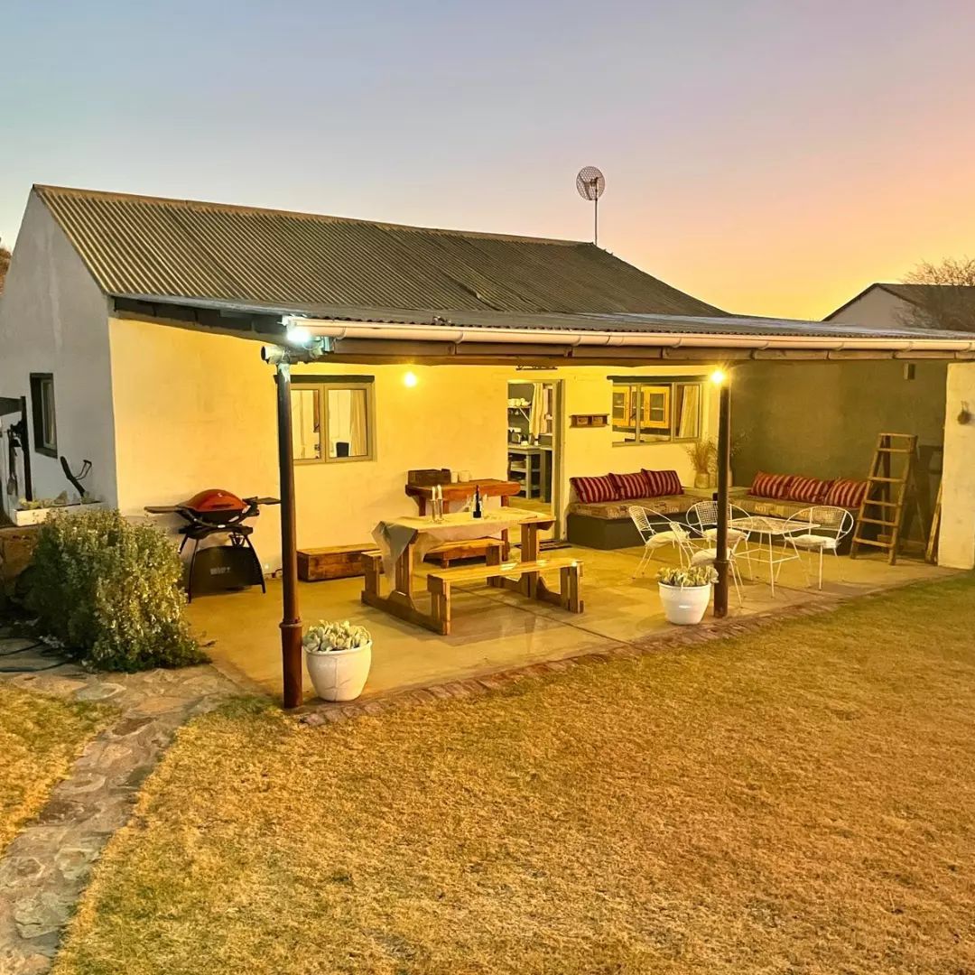 A cozy outdoor area features a wooden dining table surrounded by chairs and benches on a stone patio. A barbecue grill stands nearby, under warm lighting as the sun sets. Potted plants add a touch of greenery to the expansive lawn.