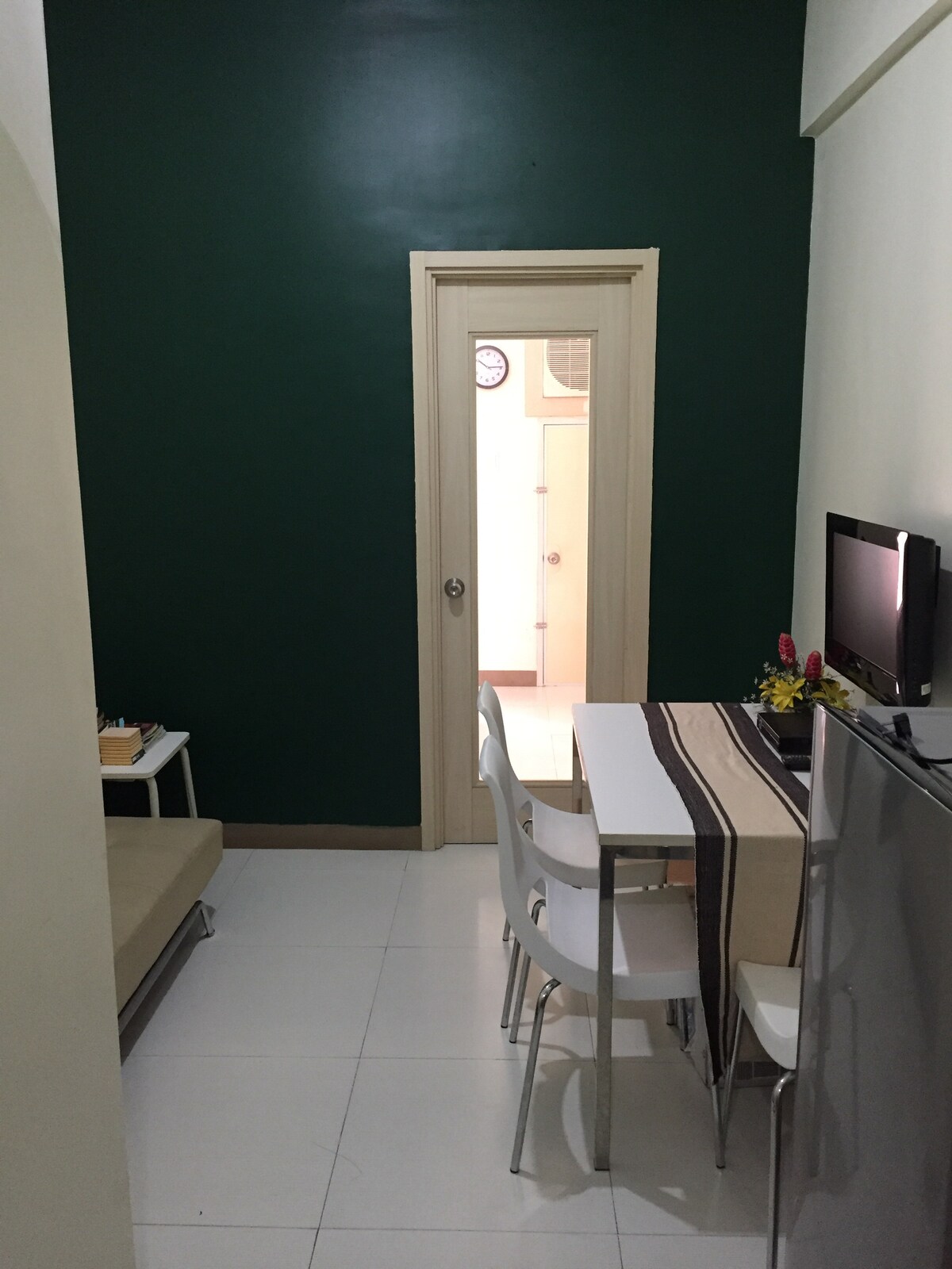 A view of the lounge area is presented, featuring a light-colored leather sofa bed alongside a small dining table with white chairs. The wall is painted a deep green, and a flat-screen TV is mounted on the nearby cabinet. A doorway is visible in the background, leading to other rooms.