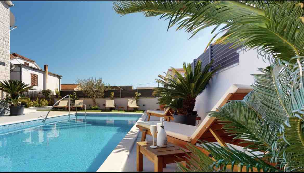 A private pool is surrounded by lush greenery and tropical plants. Sun loungers are positioned on the pool deck, while a small table holding bottle and glasses is nearby. The clear blue sky enhances the tranquil environment.