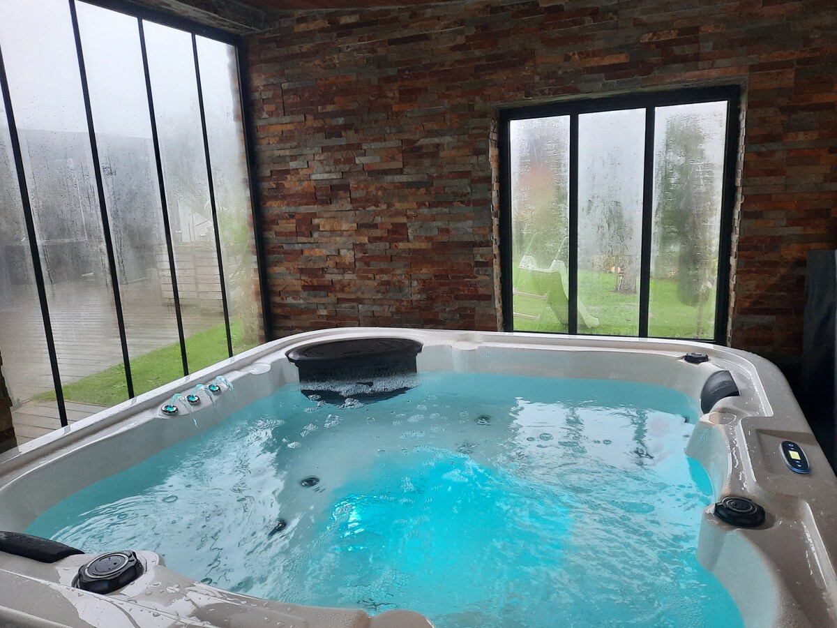 A spacious indoor spa is framed by large glass windows, showcasing natural light filtering through. The bubbling water is colored in soft blue hues, and the stone walls provide a rustic contrast to the tranquility of the area. Greenery is subtly visible through the glass.
