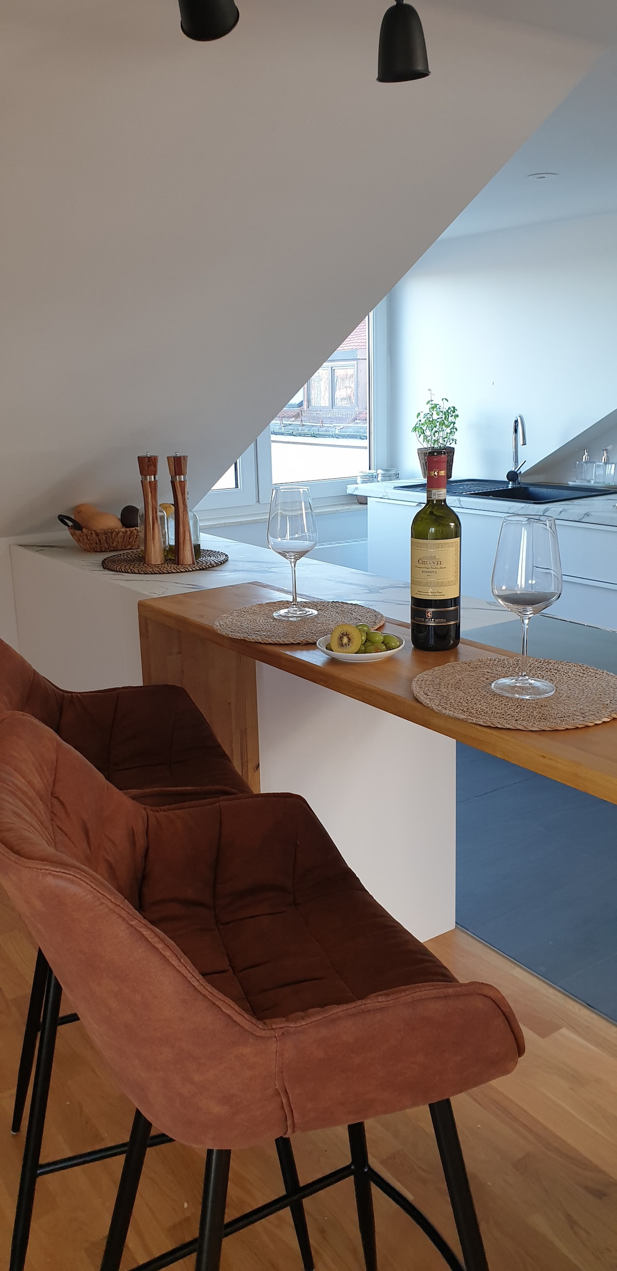 A modern kitchen bar area features two upholstered brown chairs and a wooden countertop. A bottle of wine and two glasses are displayed beside a plate with fresh fruit. The space is illuminated by natural light streaming through a triangular window.