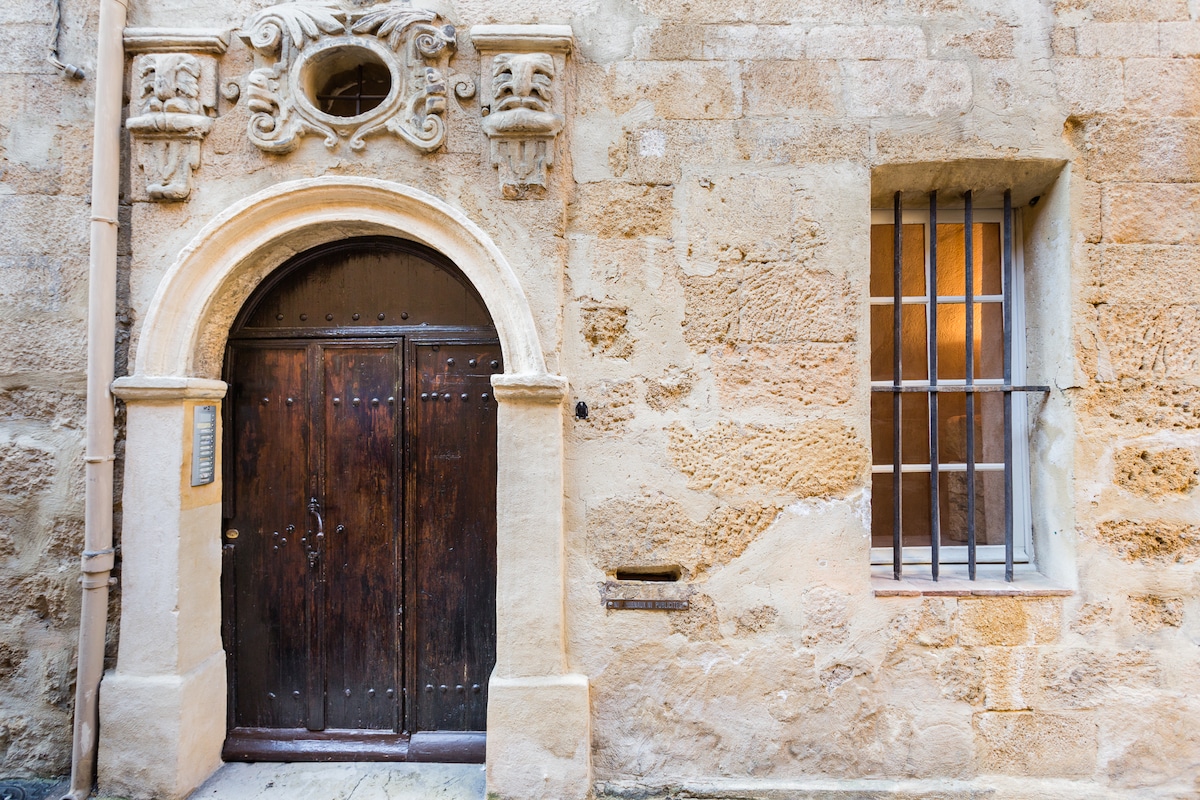 A historic entrance features an ornate wooden door framed by a light-colored stone façade. Adjacent, a barred window adds to the architectural charm, showcasing textured stone walls and subtle variations in color that reflect the age of the building.