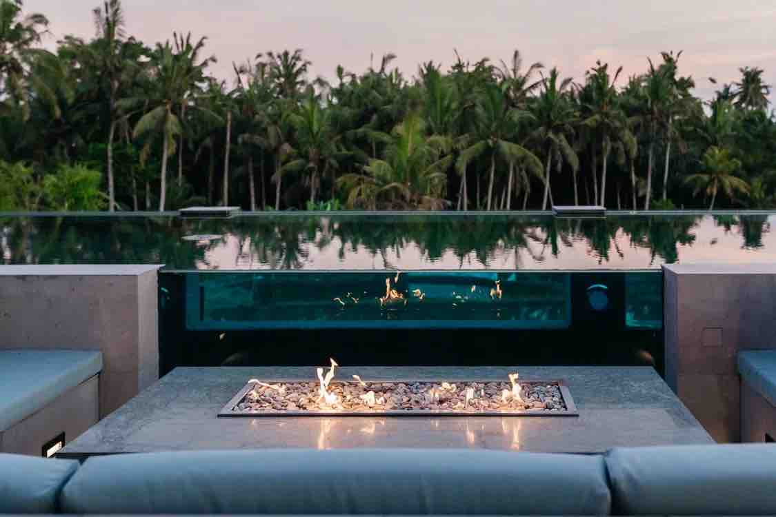 A cozy outdoor seating area features a fire pit set into a stone table, surrounded by comfortable seating. A stunning infinity pool reflects the natural landscape, with lush greenery visible beyond the water's edge.