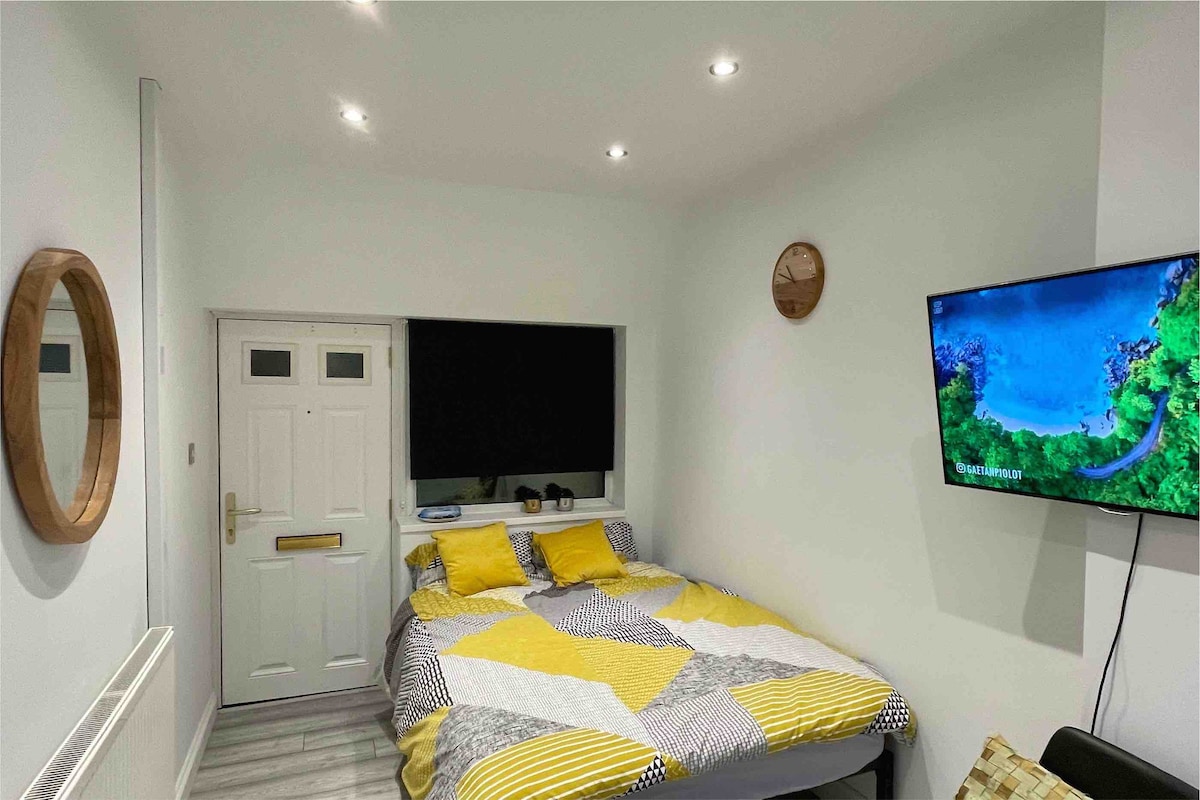 A bright and modern room features a spacious bed adorned with a geometric patterned duvet. A large flat-screen TV mounted on the wall offers entertainment options. A round mirror and a wall clock add functional elements to the space, while natural light fills the room.