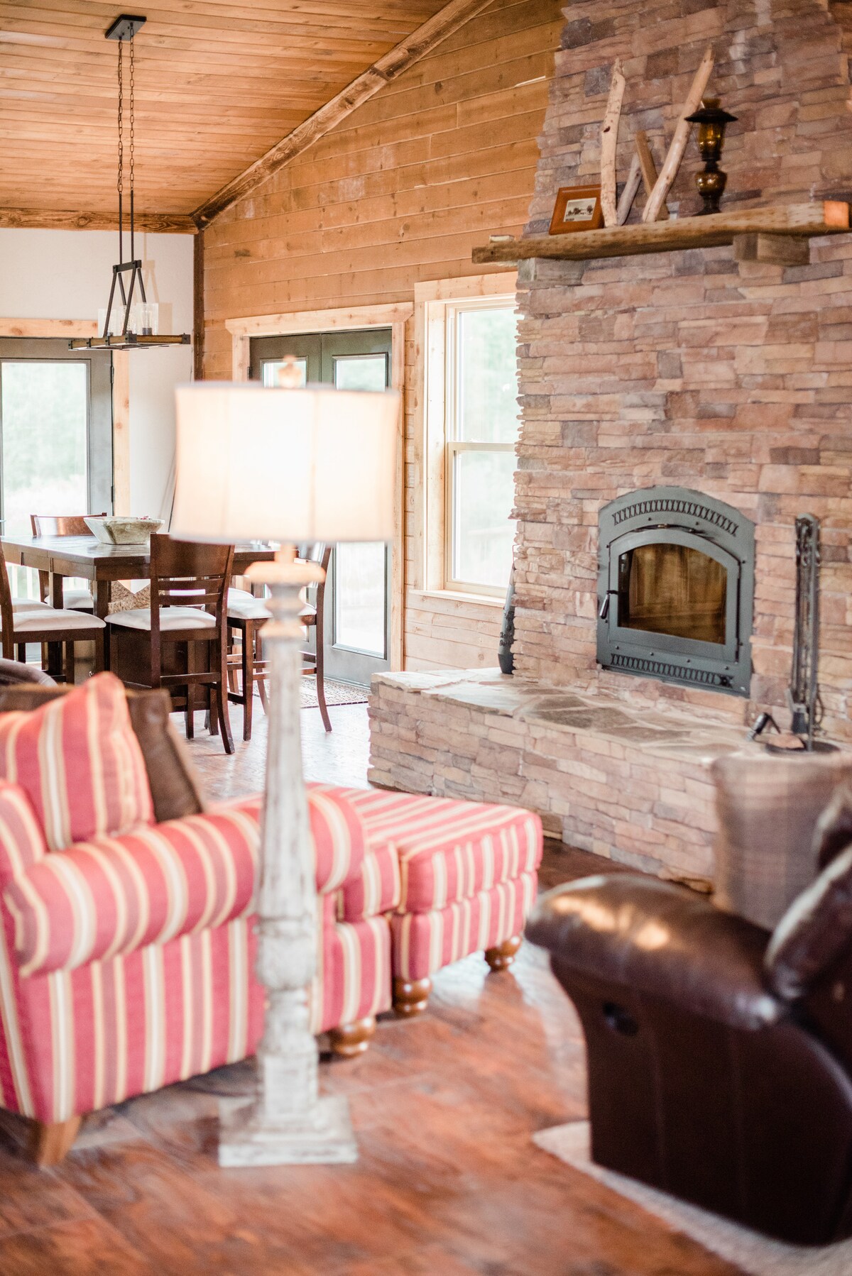 A warm living area showcases a stone fireplace set against natural wood paneling. Comfortable seating includes striped accent chairs and a leather couch. A dining table is visible in the background, illuminated by natural light streaming through large windows.