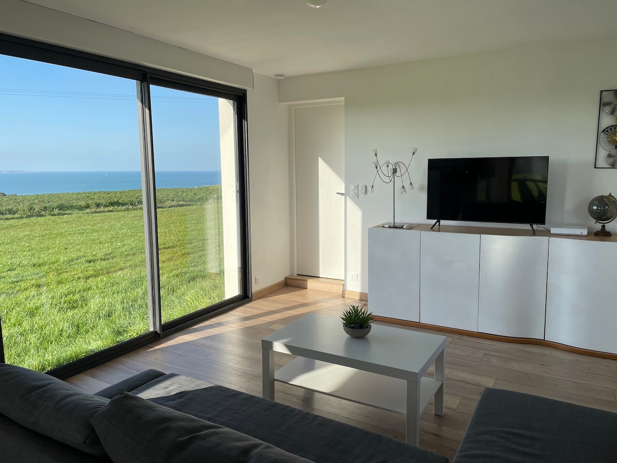 A bright living area features a large window with a view of the green landscape and sea. A modern television is positioned on a white cabinet, and a light coffee table sits atop a wooden floor. A small plant adds a touch of decor.