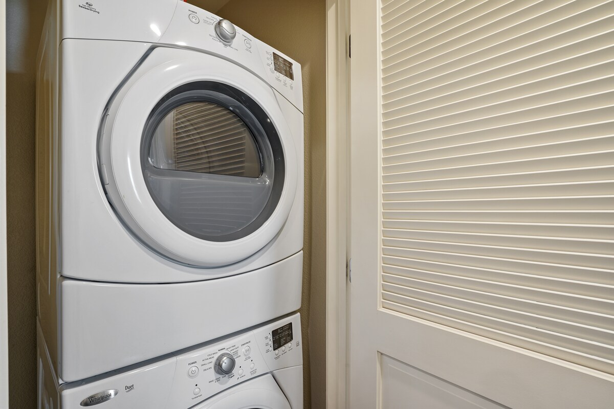 A stackable washer and dryer unit is positioned in a compact laundry area. The white appliances are neatly organized, featuring digital control panels. A closed door with horizontal slats provides a sense of privacy to the functional space.