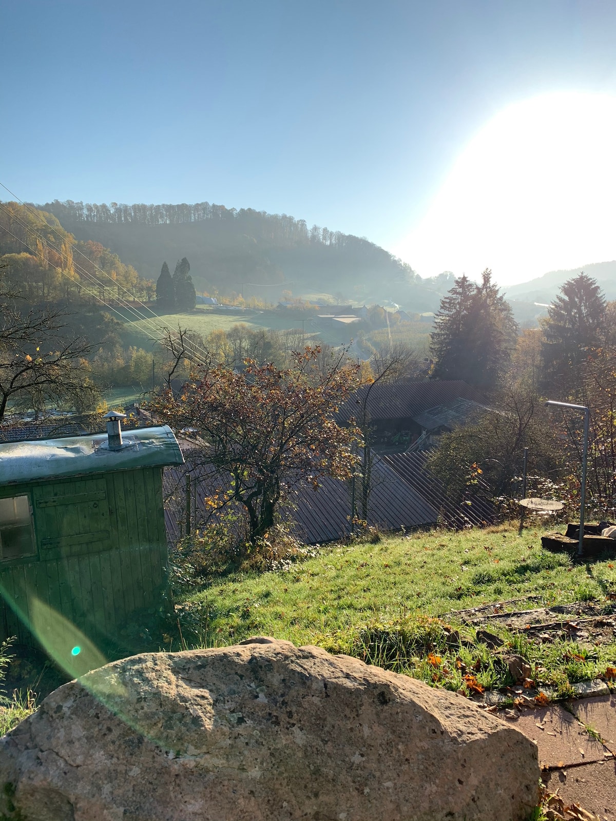 A scenic view captures a rolling landscape bathed in morning light, featuring lush greenery and distant mountains. A small shed stands in the foreground, complemented by trees adorned with autumn foliage. The soft glow of sunlight creates a serene atmosphere.