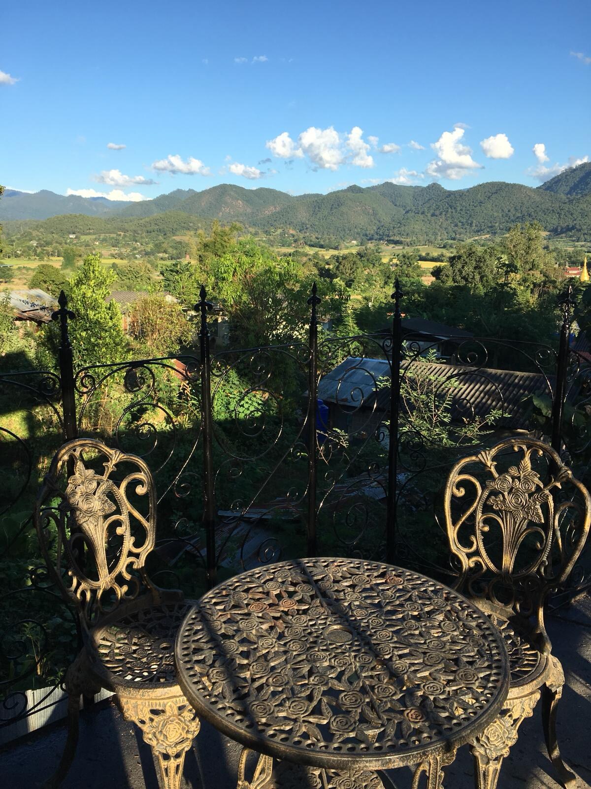 An intricately designed metal table and two matching chairs are positioned on a balcony. The scene is framed by a decorative railing, with lush greenery and rolling mountains visible in the background under a bright blue sky.