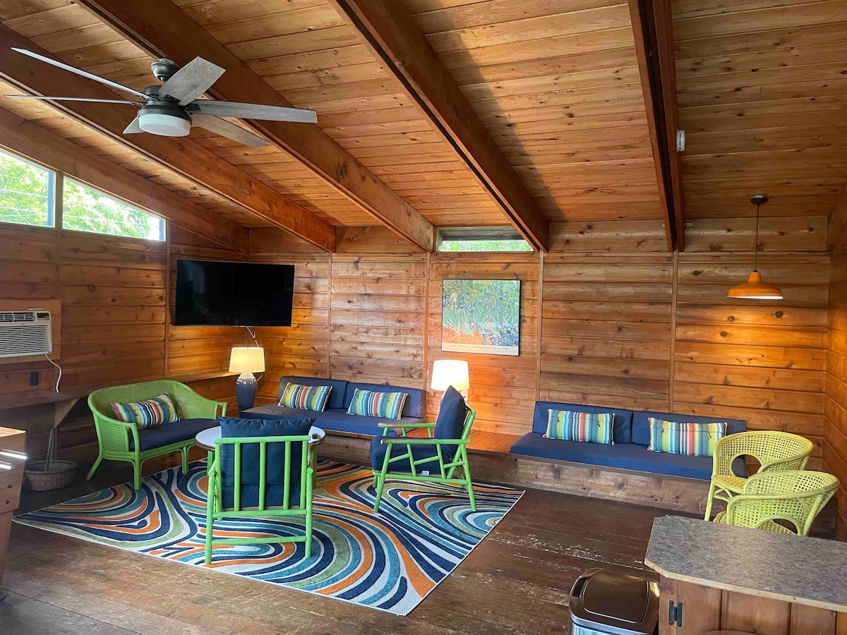 The living area features natural wood paneling and a high ceiling, with a mix of seating options in vibrant colors. A large area rug with a swirling pattern grounds the space. A flat-screen TV is mounted on the wall, complemented by bright lighting.