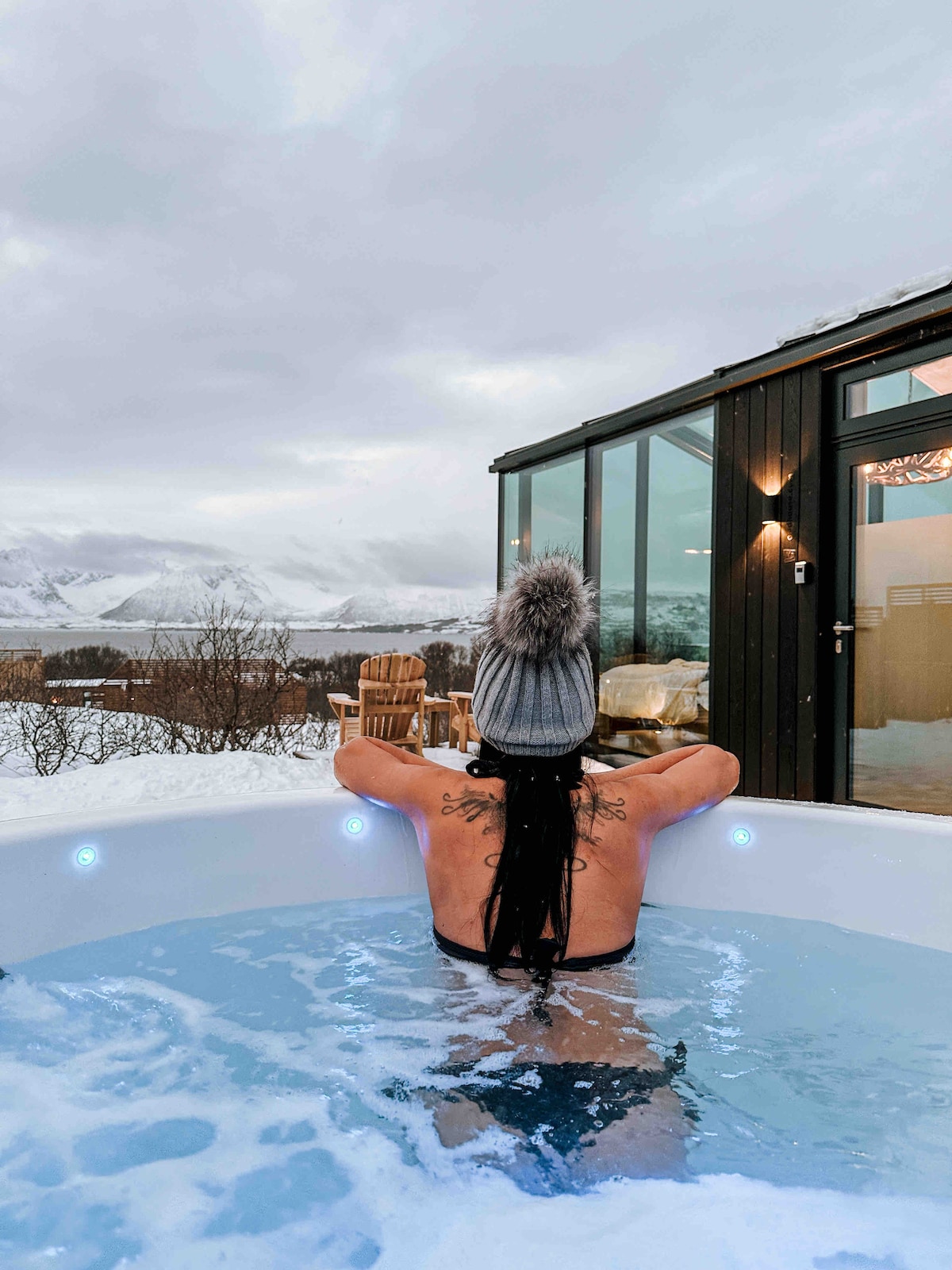 A relaxing jacuzzi is viewed from behind as a person enjoys the warm water. Snow-covered mountains are visible in the distance, while nearby wooden chairs are positioned outside the cabin. The structure's large glass walls emphasize the connection to the natural landscape.